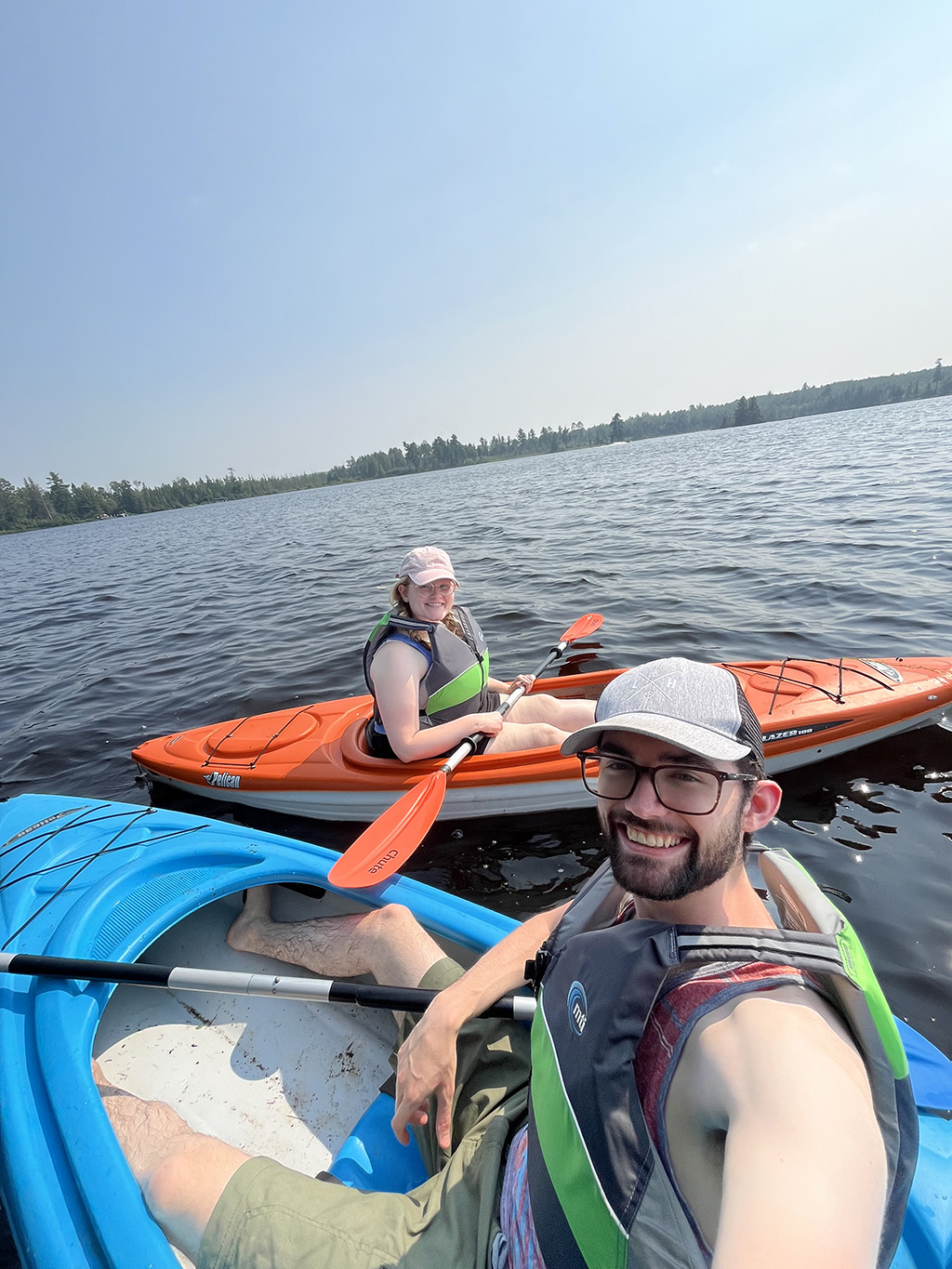 Two kayakers smile at the camera on a calm lake under a clear blue sky, one in an orange kayak and one in a blue kayak.